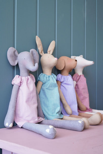 Four soft fabric animal dolls sitting side by side on a pink bench against a blue-gray paneled wall. The dolls resemble an elephant, rabbit, dog, and cat, each wearing a different pastel dress in pink, light blue, lavender, and magenta.
Photo by Tildafabrics.com [additional_image_link]
