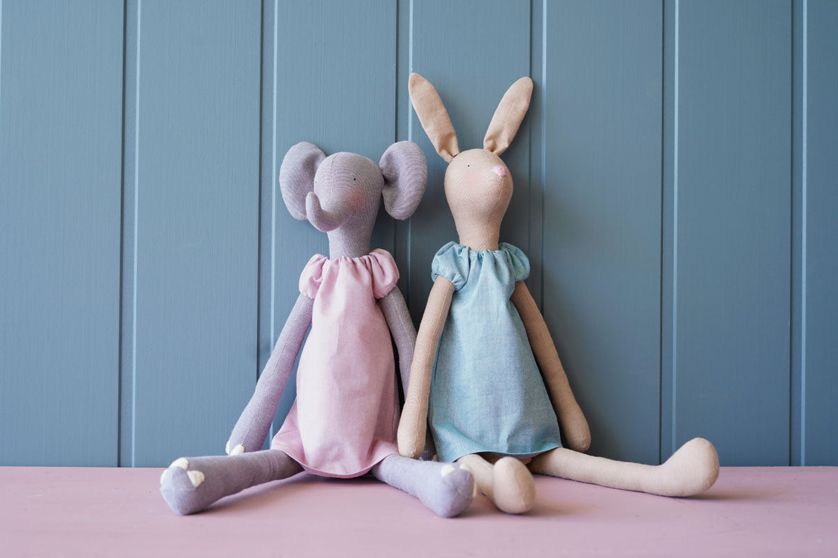 Two handmade fabric animal dolls, a pink elephant and a light blue bunny, sitting side by side against a blue paneled wall on a pink surface.
Photo by Tildafabrics.com [additional_image_link]