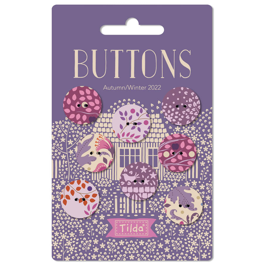 A set of eight decorative buttons from Tilda, themed 'Buttons Autumn/Winter 2022', displayed on their original purple backing card. The buttons feature whimsical, cute illustrations of houses, trees, and animals in shades of purple, pink, orange, and cream.
Photo by Tildafabrics.com [image_link]