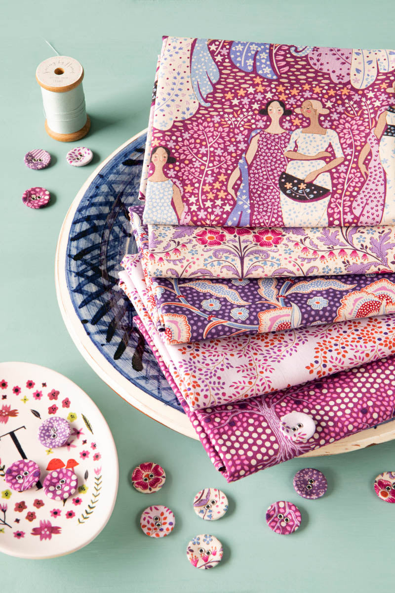 A stack of folded patterned fabric pieces in pink, purple, and white tones, arranged on a blue-and-white plate with decorative buttons, thread, and a small dish on a mint green background.
Photo by Tildafabrics.com [additional_image_link]