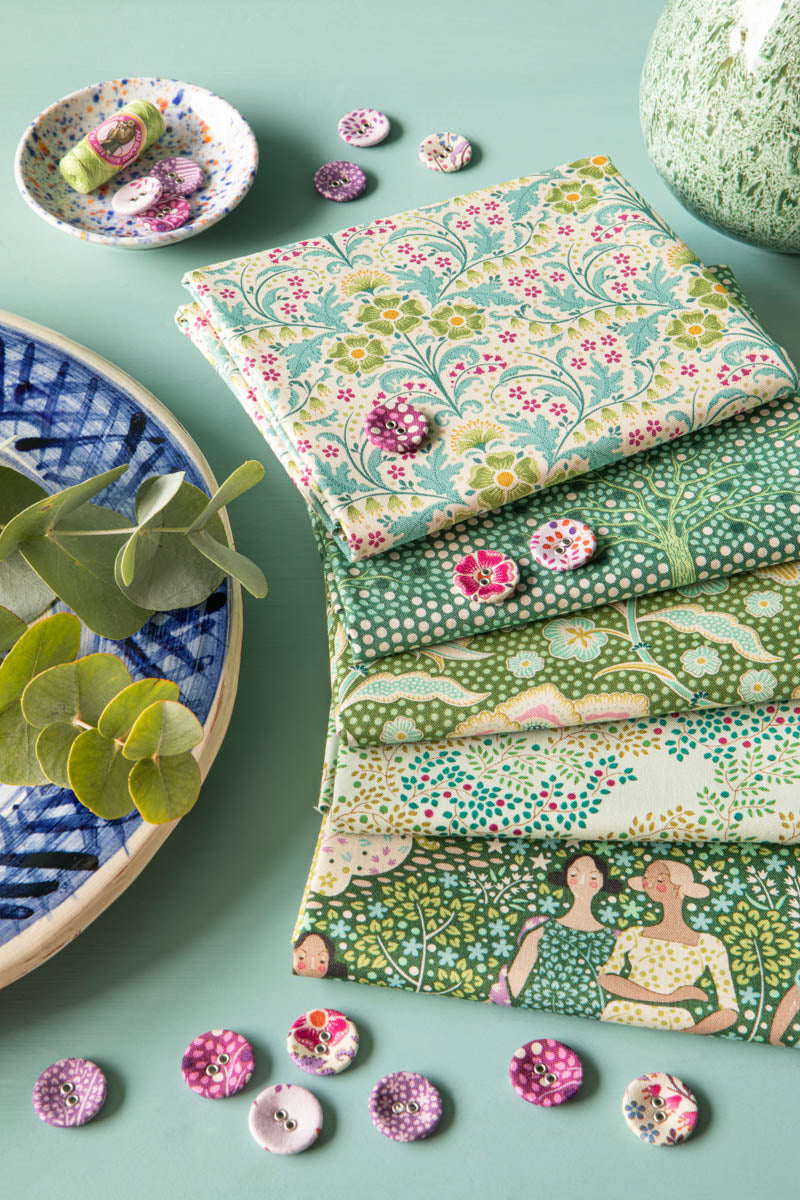 Four folded fabric pieces with floral and botanical prints in green, pink, and cream tones are arranged on a mint green surface, styled with decorative buttons, a blue patterned bowl, and green leaves.
Photo by Tildafabrics.com [additional_image_link]