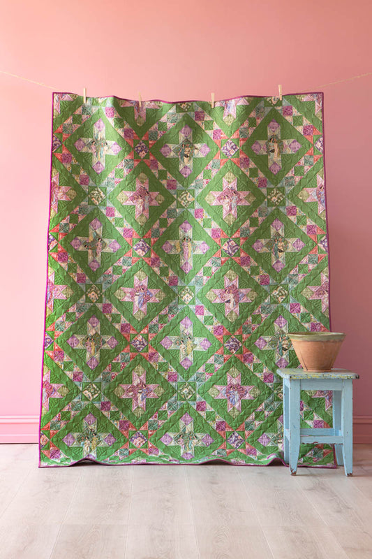 A green quilted fabric panel with a colorful floral and geometric patchwork pattern, hanging against a pink wall next to a small light blue stool with a bowl on it.
Photo by Tildafabrics.com [image_link]