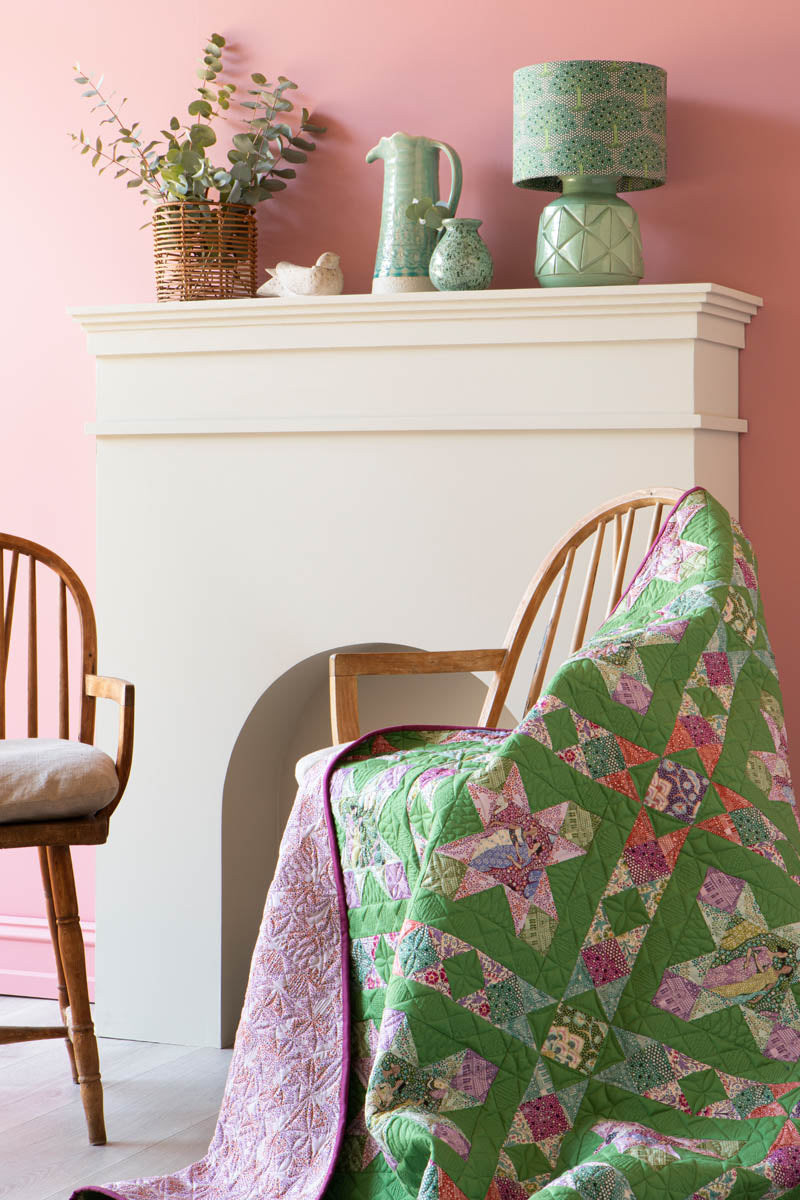 A green and pink patchwork quilt draped over wooden chairs in front of a white fireplace mantel, with a pink wall and decorative ceramic items in the background.
Photo by Tildafabrics.com [additional_image_link]