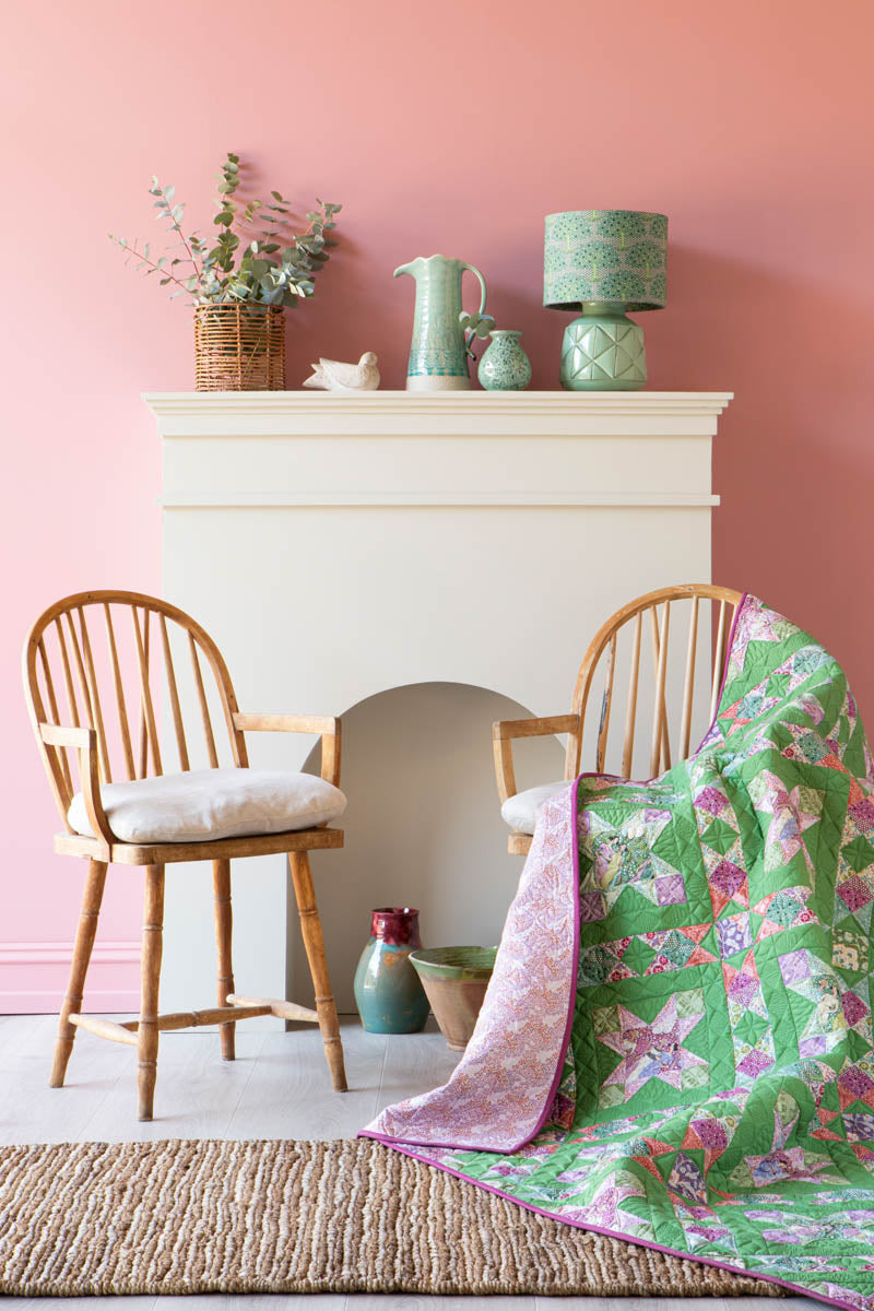 A green and pink floral quilted throw blanket is draped over a wooden spindle-back chair in a styled room with a pink wall, white fireplace, and another matching chair.
Photo by Tildafabrics.com [additional_image_link]