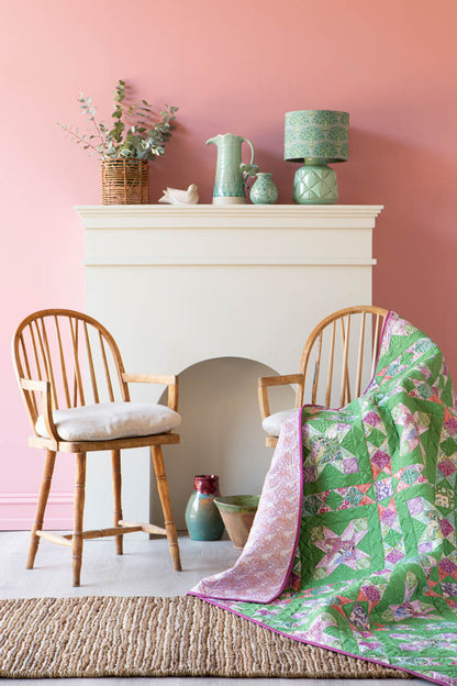 A green and pink floral quilted throw blanket is draped over a wooden spindle-back chair in a styled room with a pink wall, white fireplace, and another matching chair.
Photo by Tildafabrics.com [additional_image_link]