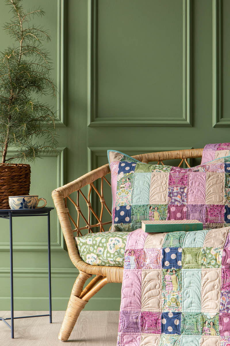 A cozy wicker armchair in a styled room with a green paneled wall, featuring a colorful patchwork quilt and matching pillow, with a small side table and potted plant nearby.
Photo by Tildafabrics.com [additional_image_link]