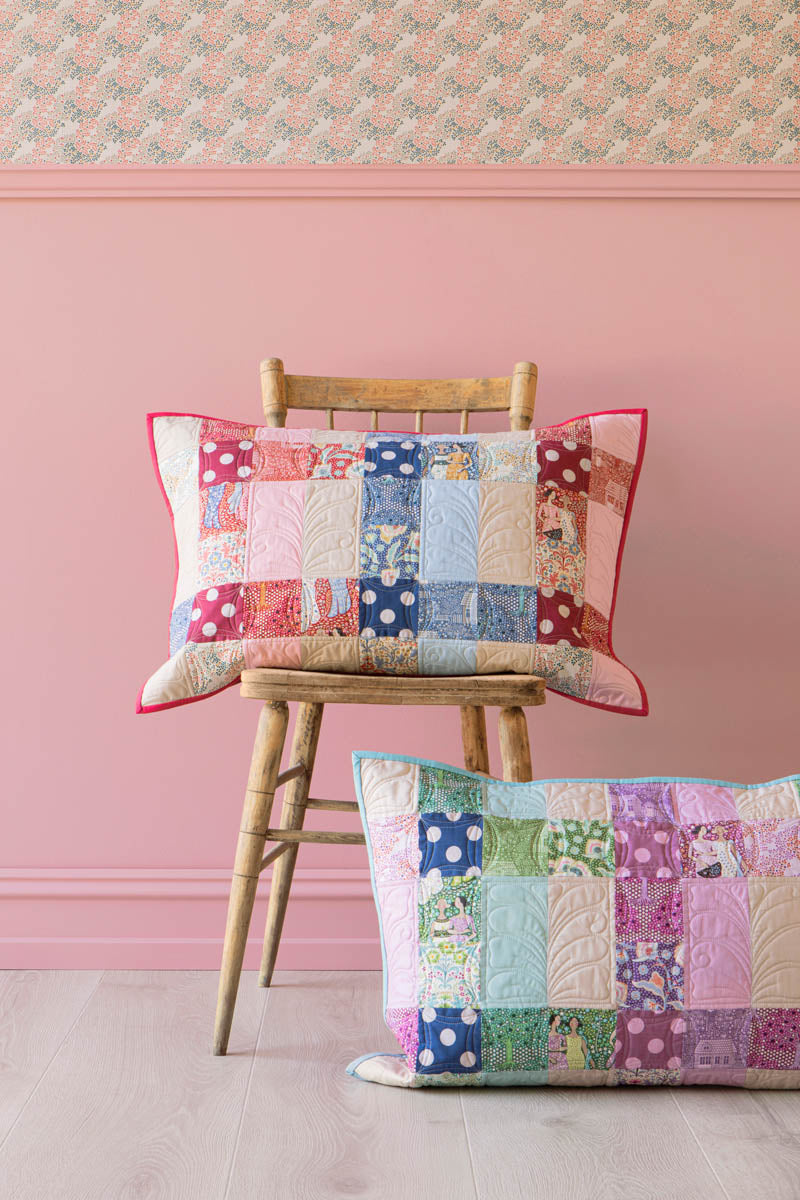 Two rectangular patchwork quilted throw pillows with red piped edges are displayed in a styled room setting. One pillow is propped up on a wooden chair, and the other lies on the floor against a pink wall with patterned wallpaper.
Photo by Tildafabrics.com [additional_image_link]