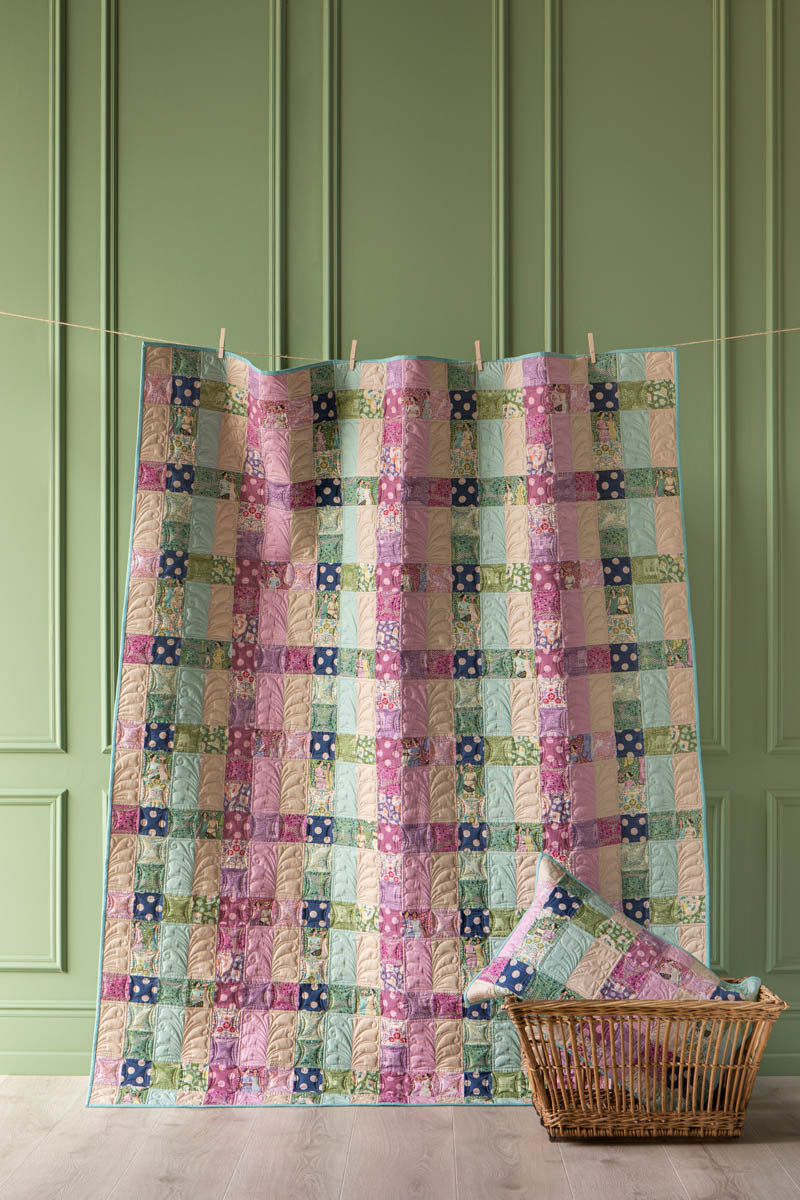 A colorful patchwork quilt hanging on a clothesline against a green paneled wall, with a matching pillow and a wicker basket on the floor.
Photo by Tildafabrics.com [additional_image_link]