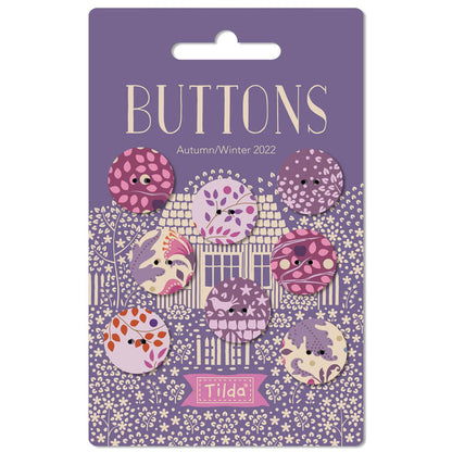 A set of eight decorative buttons from Tilda, themed 'Buttons Autumn/Winter 2022', displayed on their original purple backing card. The buttons feature whimsical, cute illustrations of houses, trees, and animals in shades of purple, pink, orange, and cream.
Photo by Tildafabrics.com [additional_image_link]