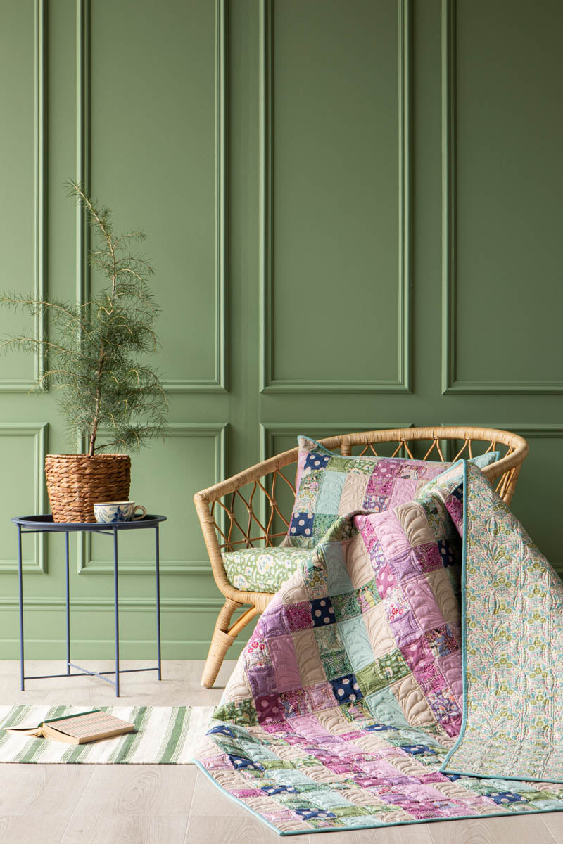 A colorful patchwork quilt draped over a wicker armchair in a styled room with green paneled walls, a small side table with a potted plant, and a striped rug.
Photo by Tildafabrics.com [additional_image_link]