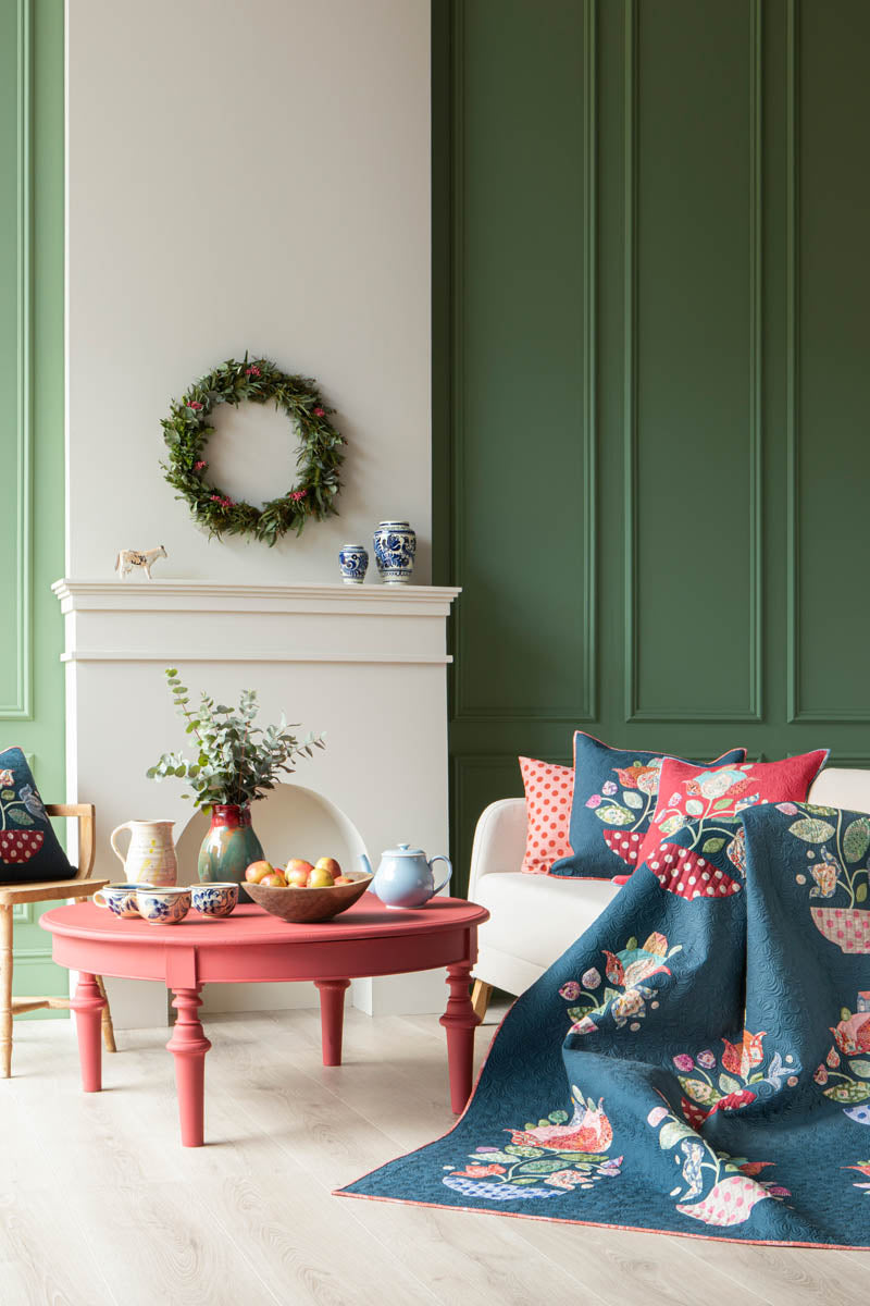 A styled living room scene featuring a navy blue floral quilt draped over a white sofa, a matching navy blue patterned area rug on the floor, and a coral red round wooden coffee table in front of a white fireplace.
Photo by Tildafabrics.com [additional_image_link]