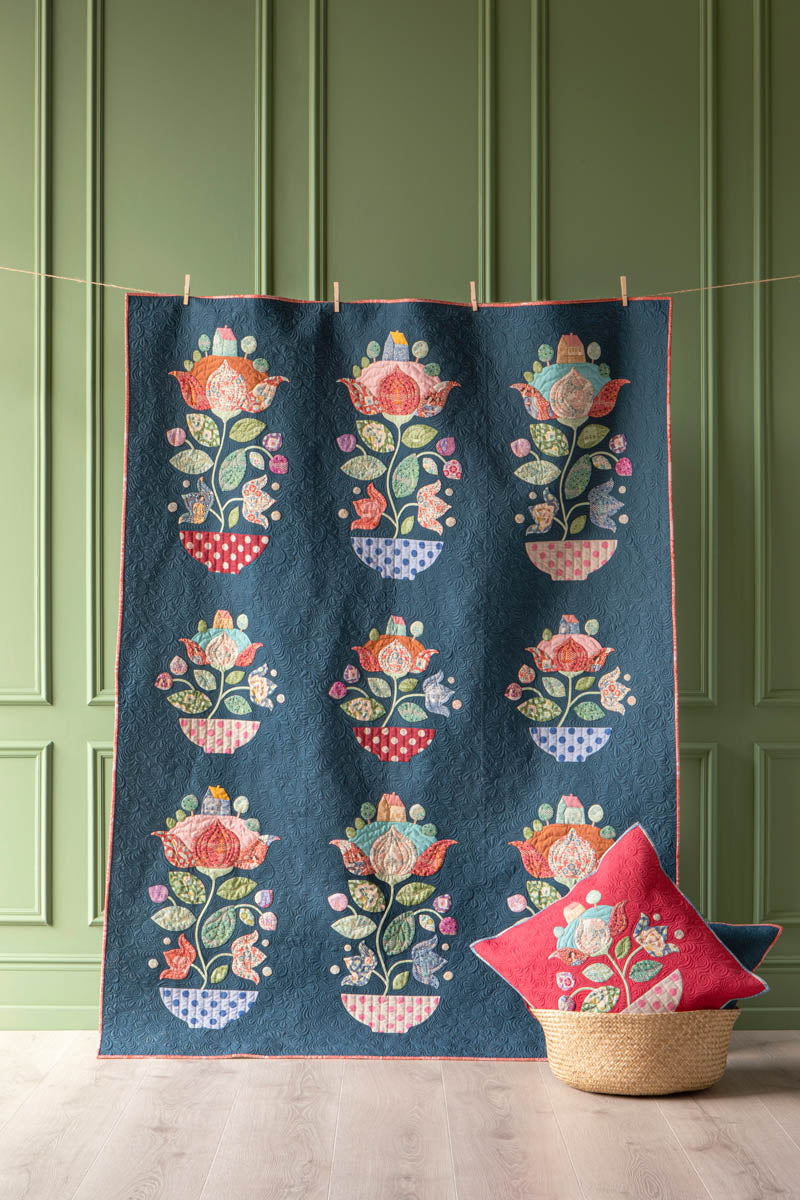 A dark navy blue quilt with a repeating floral and bowl pattern in pink, orange, green, and cream, hanging on a string against a green paneled wall with a woven basket and red pillow at the bottom.
Photo by Tildafabrics.com [additional_image_link]