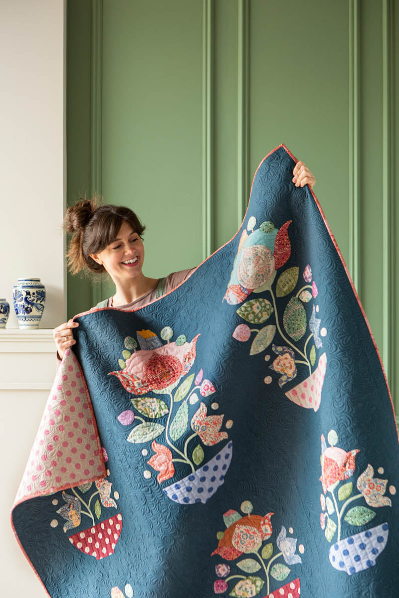 A woman holds up a large quilted blanket featuring a colorful bird and floral pattern on a dark blue background, with a pink polka-dot reverse side visible on one corner. She is standing indoors against a green paneled wall.
Photo by Tildafabrics.com [additional_image_link]