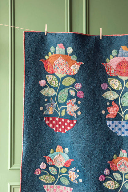 A quilt with a dark blue background and colorful floral and bird motifs, hanging from a string with wooden clothespins against a green paneled door.
Photo by Tildafabrics.com [additional_image_link]