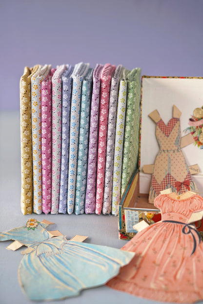 A set of vintage doll clothes and matching fabric dress-up books arranged on a light blue surface against a lavender background. The books are stacked vertically, showing various pastel floral patterns in pink, blue, green, and yellow.
Photo by Tildafabrics.com [additional_image_link]