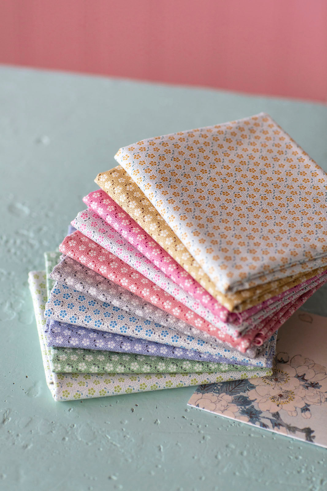 A stack of folded patterned fabric squares in soft pastel colors, including cream, yellow, pink, purple, and green, arranged on a light gray surface against a pink background.
Photo by Tildafabrics.com [additional_image_link]