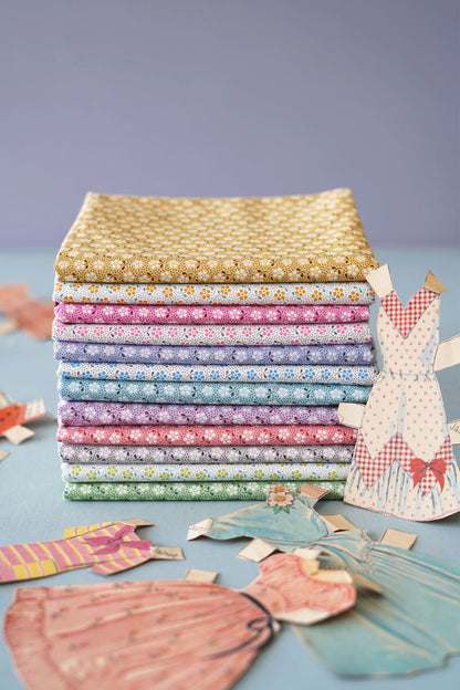 A stack of colorful patterned fabric squares in pastel and warm tones, arranged on a light blue surface with paper dress cutouts around them against a soft lavender background.
Photo by Tildafabrics.com [additional_image_link]