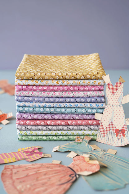 A stack of colorful patterned fabric squares in pastel and warm tones, arranged on a light blue surface with paper dress cutouts around them against a soft lavender background.
Photo by Tildafabrics.com [additional_image_link]