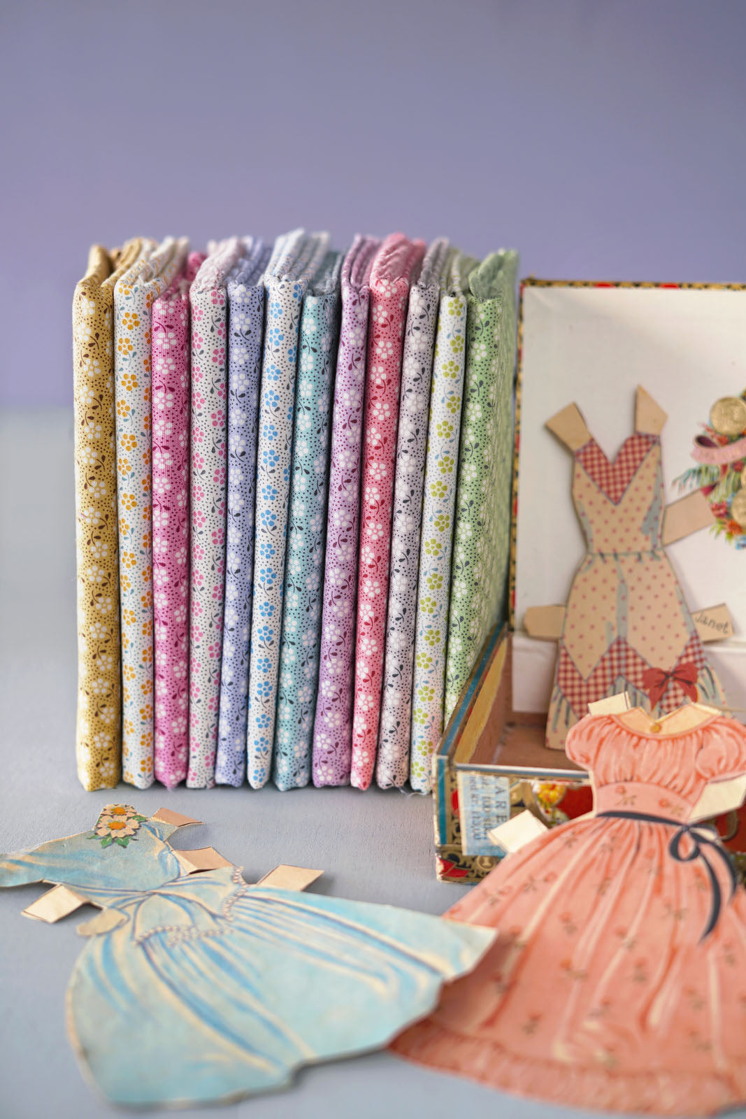 A set of vintage doll clothes and matching fabric dress-up books arranged on a light blue surface against a lavender background. The books are stacked vertically, showing various pastel floral patterns in pink, blue, green, and yellow.
Photo by Tildafabrics.com [additional_image_link]