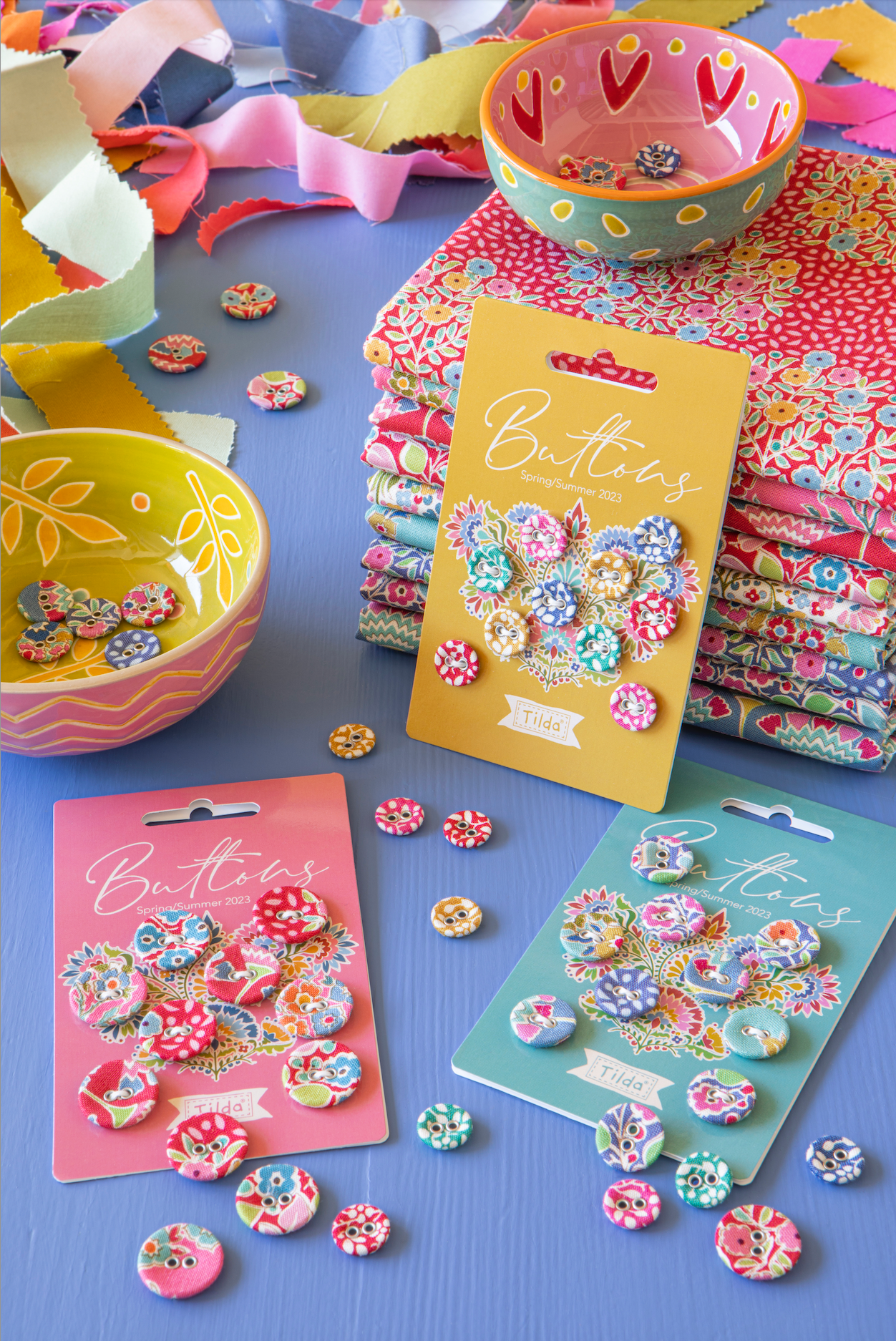 Three packages of decorative buttons in floral and patterned designs are arranged on a blue surface with matching fabric scraps, a bowl, and ribbon around them. The buttons are the product, shown in retail cards with visible 'Buttons' text.
Photo by Tildafabrics.com [additional_image_link]