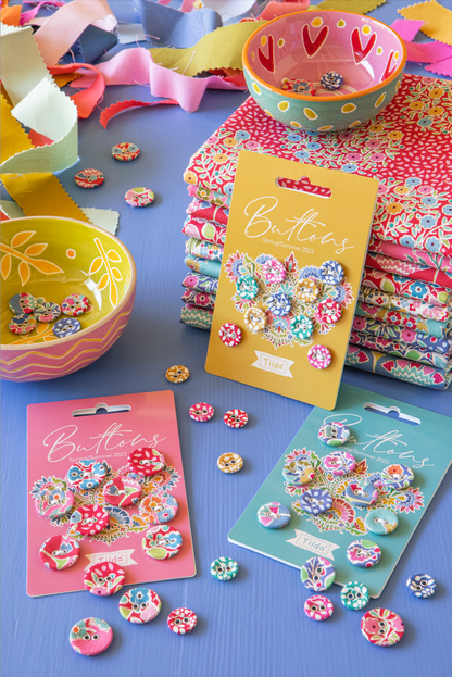 Three packages of decorative buttons in floral and patterned designs are arranged on a blue surface with matching fabric scraps, a bowl, and ribbon around them. The buttons are the product, shown in retail cards with visible 'Buttons' text.
Photo by Tildafabrics.com [additional_image_link]