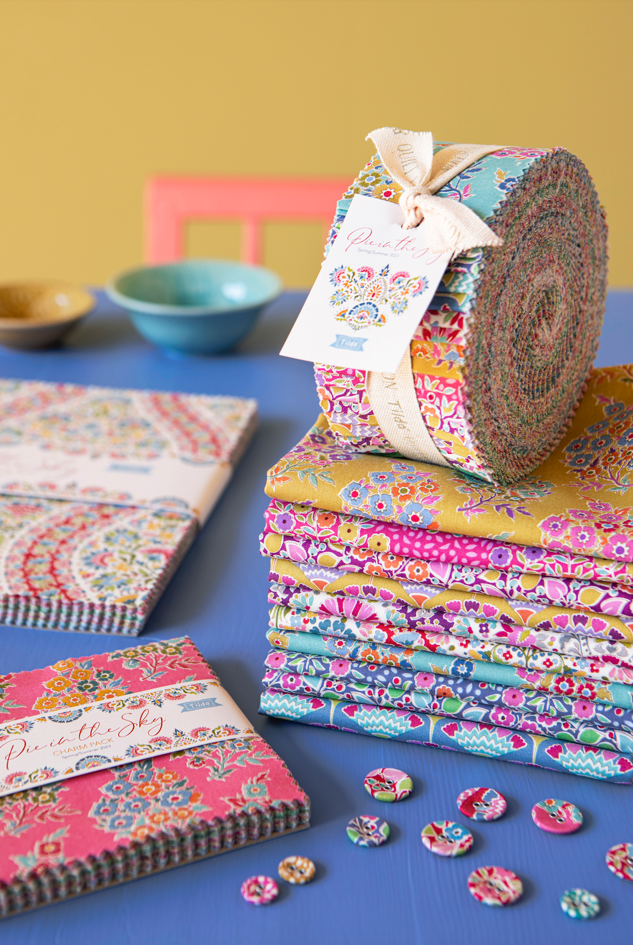 A styled product display of fabric bundles and folded fabric pieces with floral and paisley patterns, tied with cream ribbon and labeled 'Painting Shy'. The fabrics are arranged on a blue surface against a mustard yellow background, with small bowls and a pink chair visible in the blurred background.
Photo by Tildafabrics.com [additional_image_link]