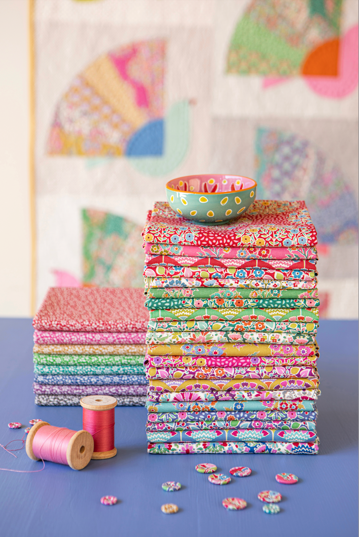Stacks of colorful patterned quilting fabrics on a blue surface, with a small patterned bowl on top of the right stack, spools of thread, loose fabric pieces, and a quilted wall hanging in the background.
Photo by Tildafabrics.com [additional_image_link]