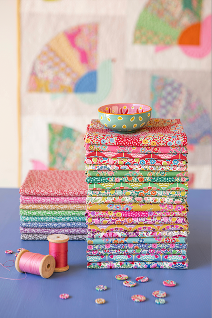 Stacks of colorful patterned quilting fabrics on a blue surface, with a small patterned bowl on top of the right stack, spools of thread, loose fabric pieces, and a quilted wall hanging in the background.
Photo by Tildafabrics.com [additional_image_link]