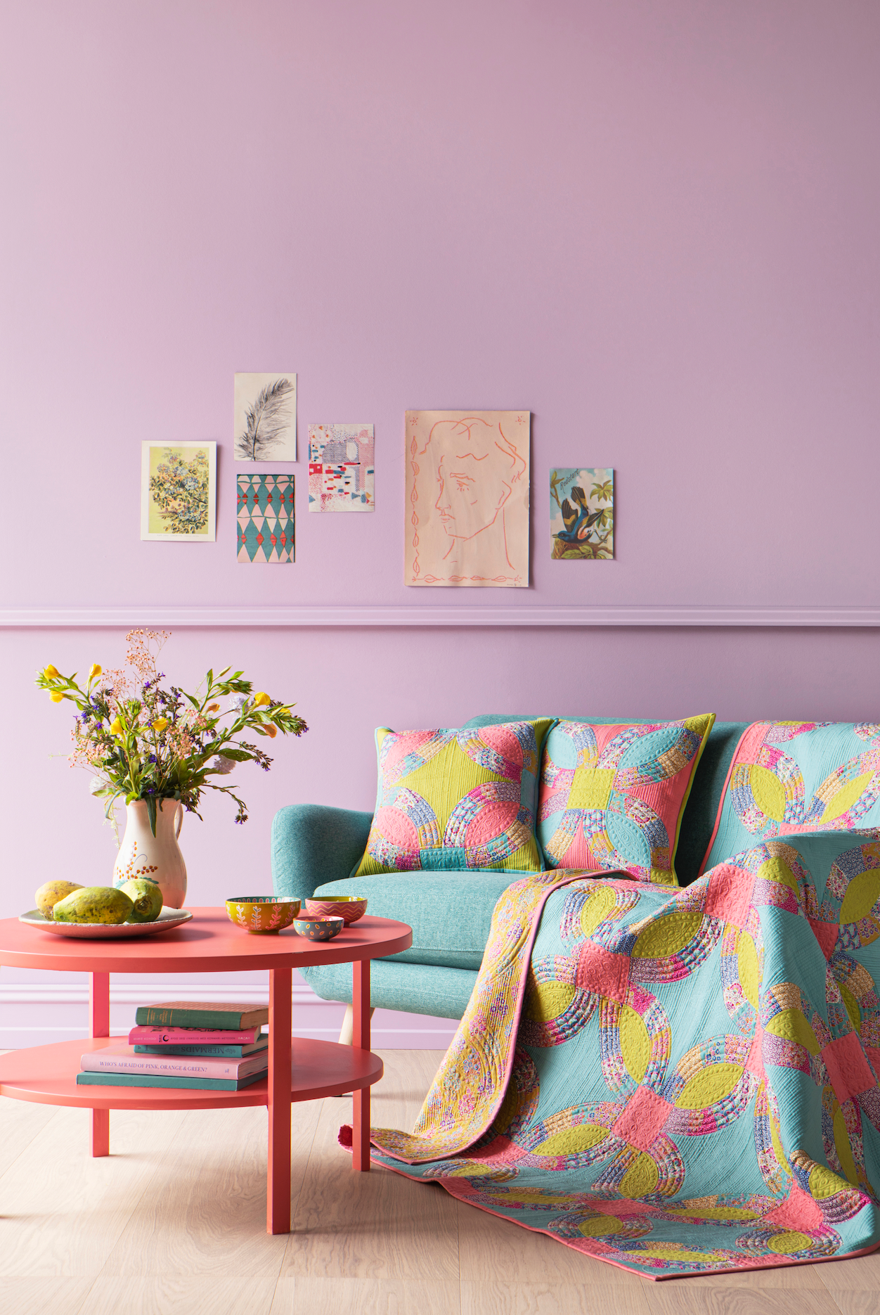 A styled interior scene with a pastel lavender wall, framed art prints, a mint green sofa, two patterned pink and yellow throw pillows, and a matching patterned quilt draped over the arm. A round pink side table with a vase of flowers and small bowls sits beside the sofa.
Photo by Tildafabrics.com [additional_image_link]