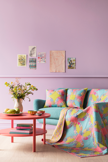 A styled interior scene with a pastel lavender wall, framed art prints, a mint green sofa, two patterned pink and yellow throw pillows, and a matching patterned quilt draped over the arm. A round pink side table with a vase of flowers and small bowls sits beside the sofa.
Photo by Tildafabrics.com [additional_image_link]