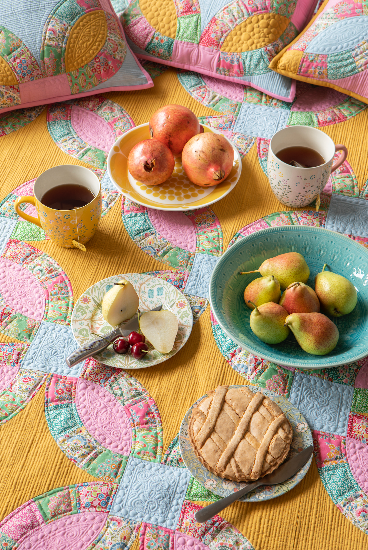 A styled tabletop scene with a colorful quilt, two mugs of tea, a plate of pomegranates, a bowl of pears, a small plate with a pear and cheese, and a tart on a matching quilted textile.
Photo by Tildafabrics.com [additional_image_link]