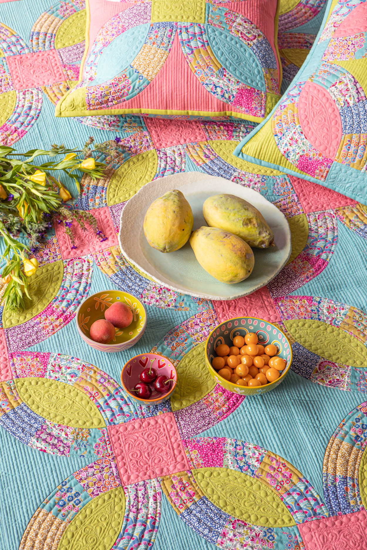 A colorful quilted textile set styled in a flat lay, featuring a quilt and matching pillows with a vibrant geometric and floral pattern in pink, yellow, and blue. The scene is styled with small bowls of fruit and flowers on a white plate.
Photo by Tildafabrics.com [additional_image_link]