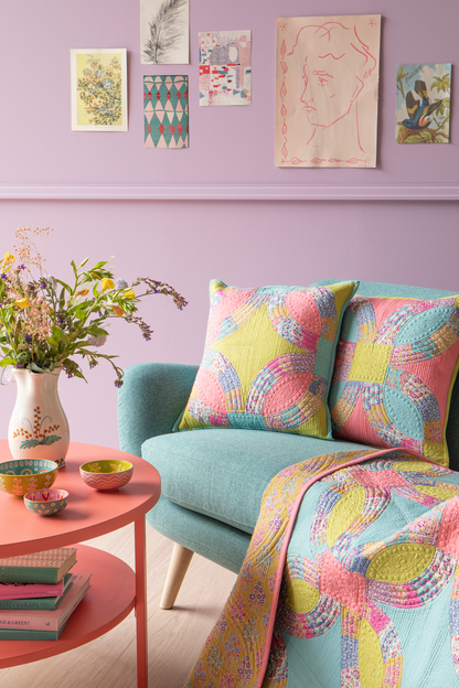 A styled interior scene with a pastel lavender wall, framed art prints, a mint green sofa, two patterned pink and yellow throw pillows, and a matching patterned quilt draped over the arm. A round pink side table with a vase of flowers and small bowls sits beside the sofa.
Photo by Tildafabrics.com [additional_image_link]