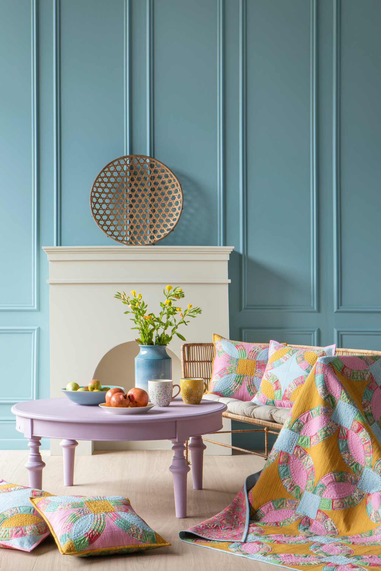 A styled interior scene with a pastel lavender round coffee table, colorful patterned cushions and quilt, a blue vase with green stems, and a white fireplace against a blue paneled wall.
Photo by Tildafabrics.com [additional_image_link]