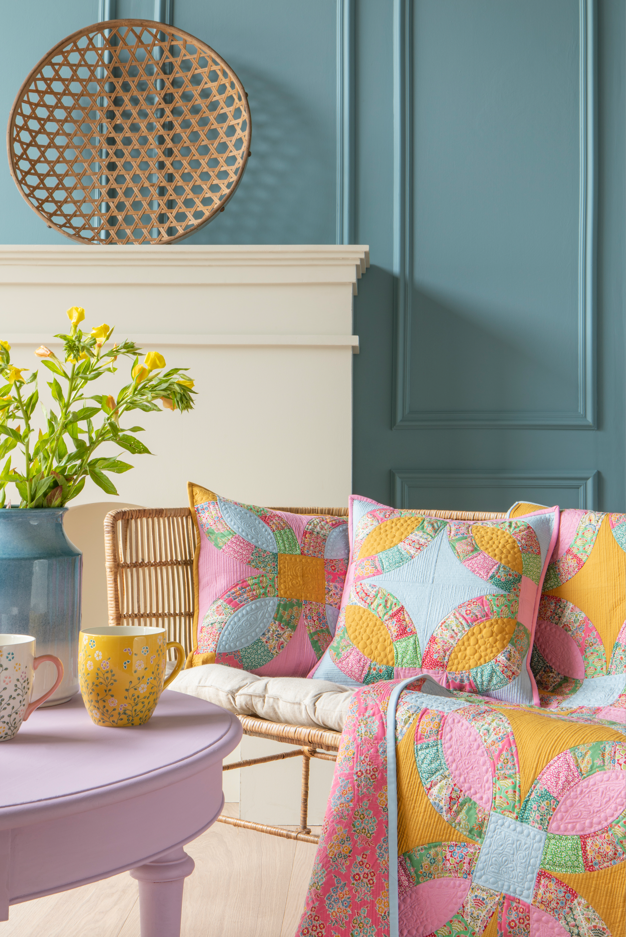 A styled interior scene showing a pastel quilted throw blanket and matching decorative pillows with a colorful butterfly patchwork pattern, arranged on a sofa in a room with blue paneled walls and a lavender side table.
Photo by Tildafabrics.com [additional_image_link]