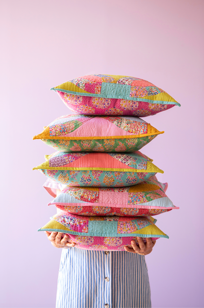A stack of six colorful patchwork pillows held up by a person wearing a blue and white striped shirt against a solid light pink background.
Photo by Tildafabrics.com [additional_image_link]
