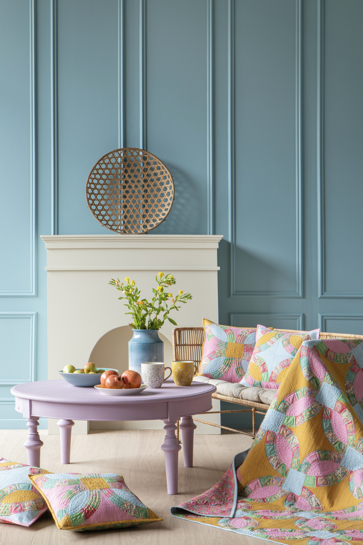 A styled interior scene with a pastel lavender round coffee table, colorful patterned cushions and quilt, a blue vase with green stems, and a white fireplace against a blue paneled wall.
Photo by Tildafabrics.com [additional_image_link]