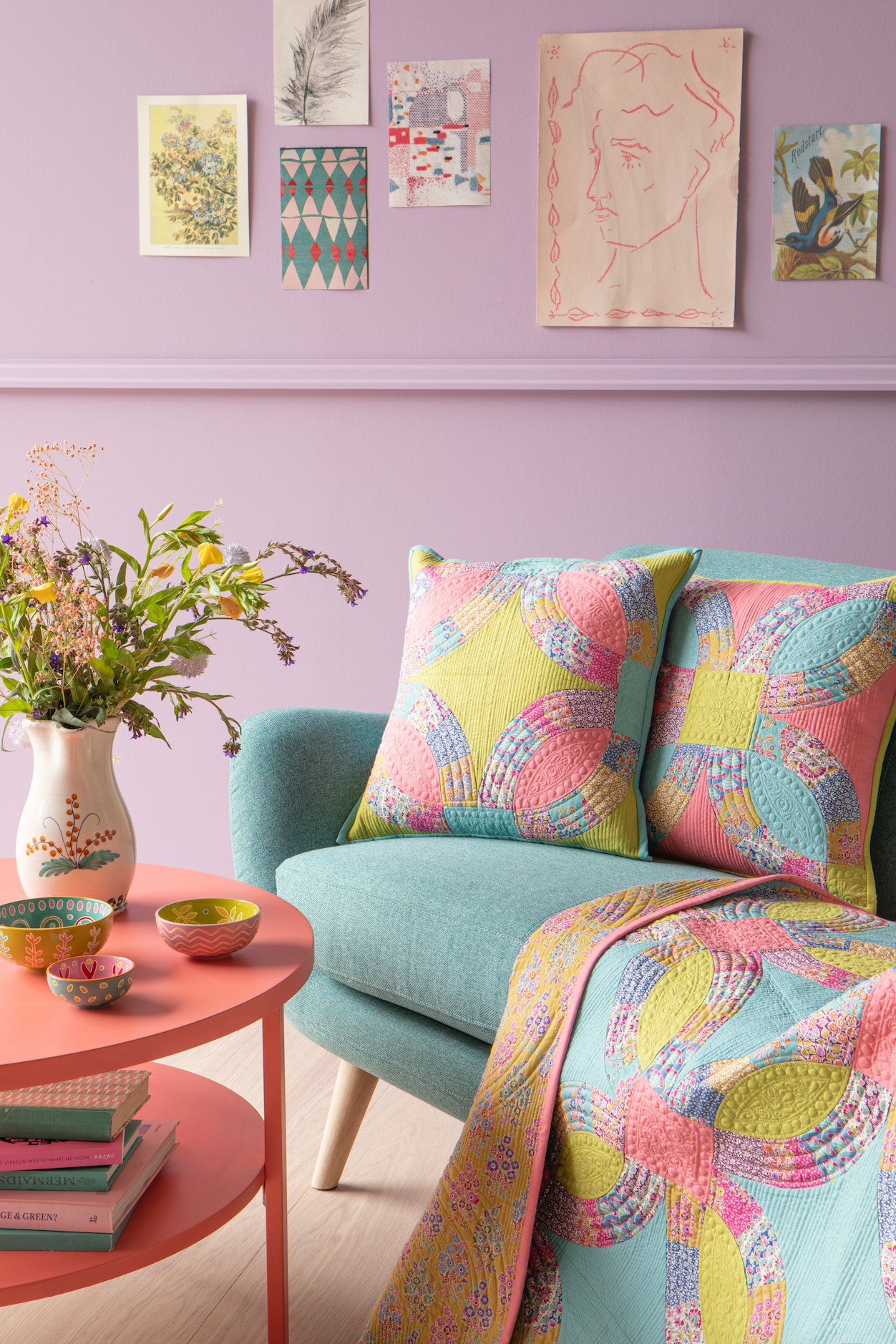 A styled interior scene with a pastel lavender wall, framed art prints, a mint green sofa, two patterned pink and yellow throw pillows, and a matching patterned blanket draped over the arm. A round pink side table with a vase of flowers and small bowls sits beside the sofa.
Photo by Tildafabrics.com [additional_image_link]