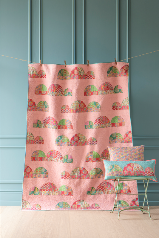 A pink quilted blanket with a repeating pattern of stylized houses in green, red, and orange, hanging from a string with wooden clips against a teal paneled wall. A small green metal stool with two patterned pillows sits on the floor next to it.
Photo by Tildafabrics.com [image_link]