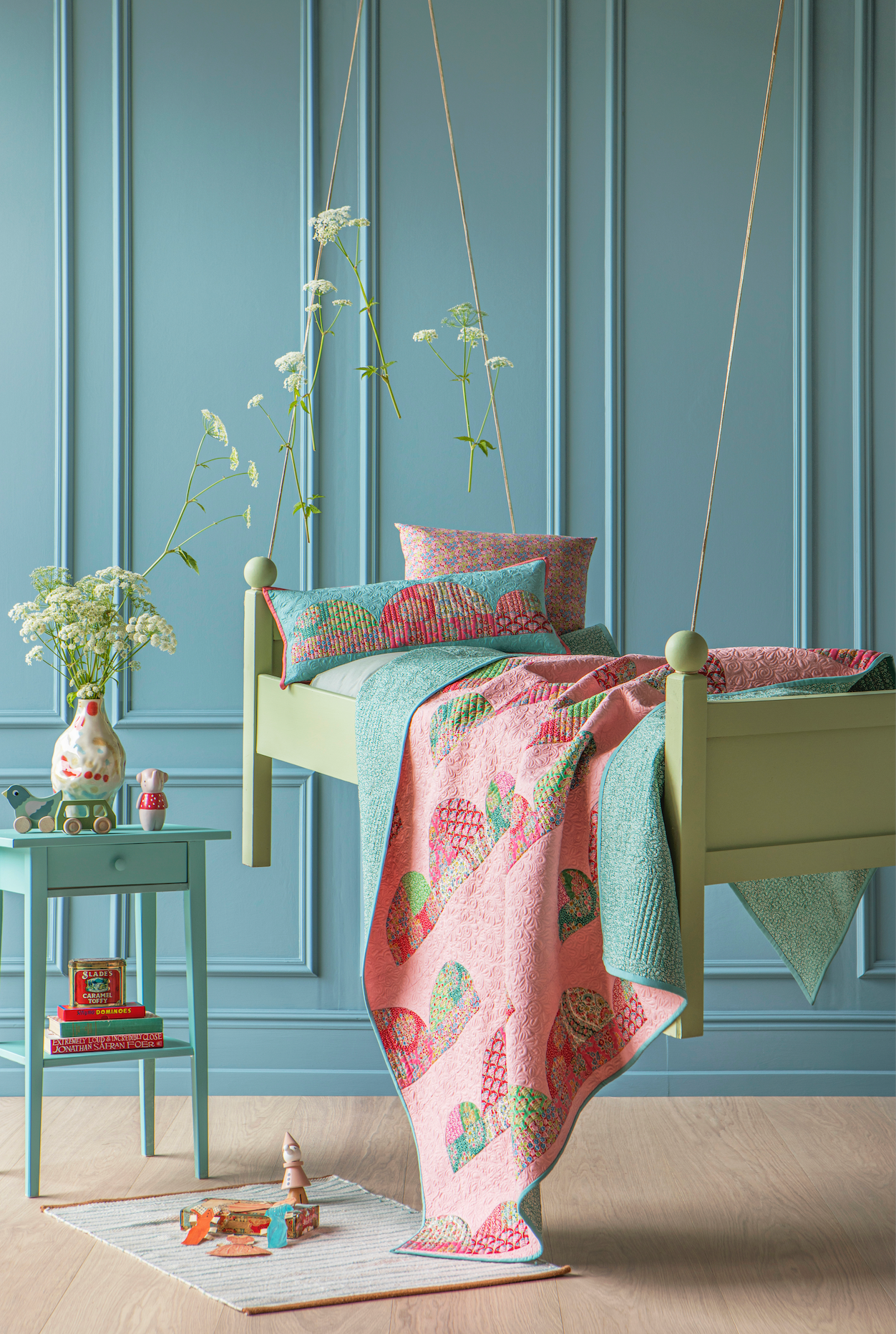 A styled children's bedroom scene with a hanging swing bed. The bed frame is light sage green, dressed in a pink quilted blanket with colorful fruit patterns and teal pillows, set against a blue paneled wall with a turquoise side table and flowers.
Photo by Tildafabrics.com [additional_image_link]