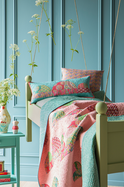 A styled bedroom scene with a light wood bed dressed in a pink quilted blanket and matching patterned pillows, set against a blue paneled wall with hanging white flowers and a turquoise side table.
Photo by Tildafabrics.com [additional_image_link]