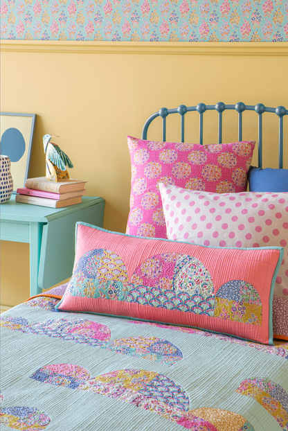 A styled bedroom scene with a bed dressed in pastel patterned bedding and several decorative pillows, including a pink striped pillow and a pink-and-white polka dot pillow, against a yellow wall and mint side table.
Photo by Tildafabrics.com [additional_image_link]