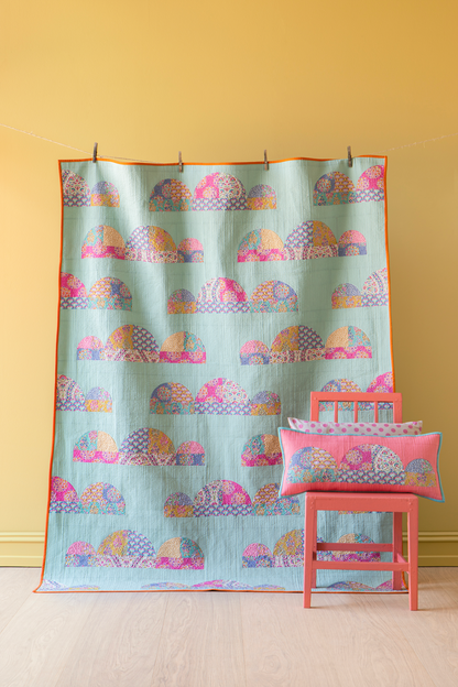 A light blue quilted fabric panel with a repeating pink, purple, and orange cloud-like pattern, hanging on a yellow wall next to a coral-pink wooden chair with matching pillows.
Photo by Tildafabrics.com [additional_image_link]