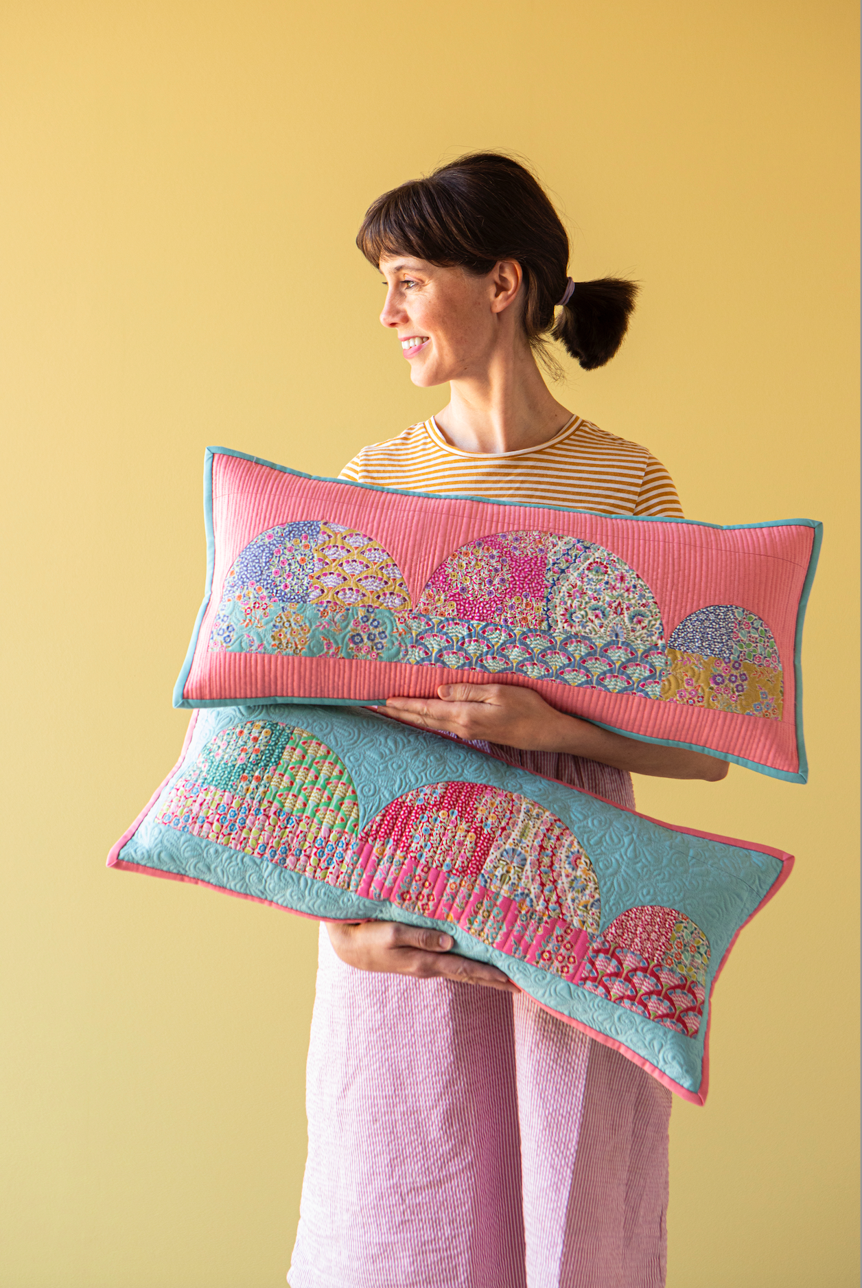 A woman holding two colorful quilted lumbar pillows with a scalloped paisley pattern, set against a solid yellow background.
Photo by Tildafabrics.com [additional_image_link]