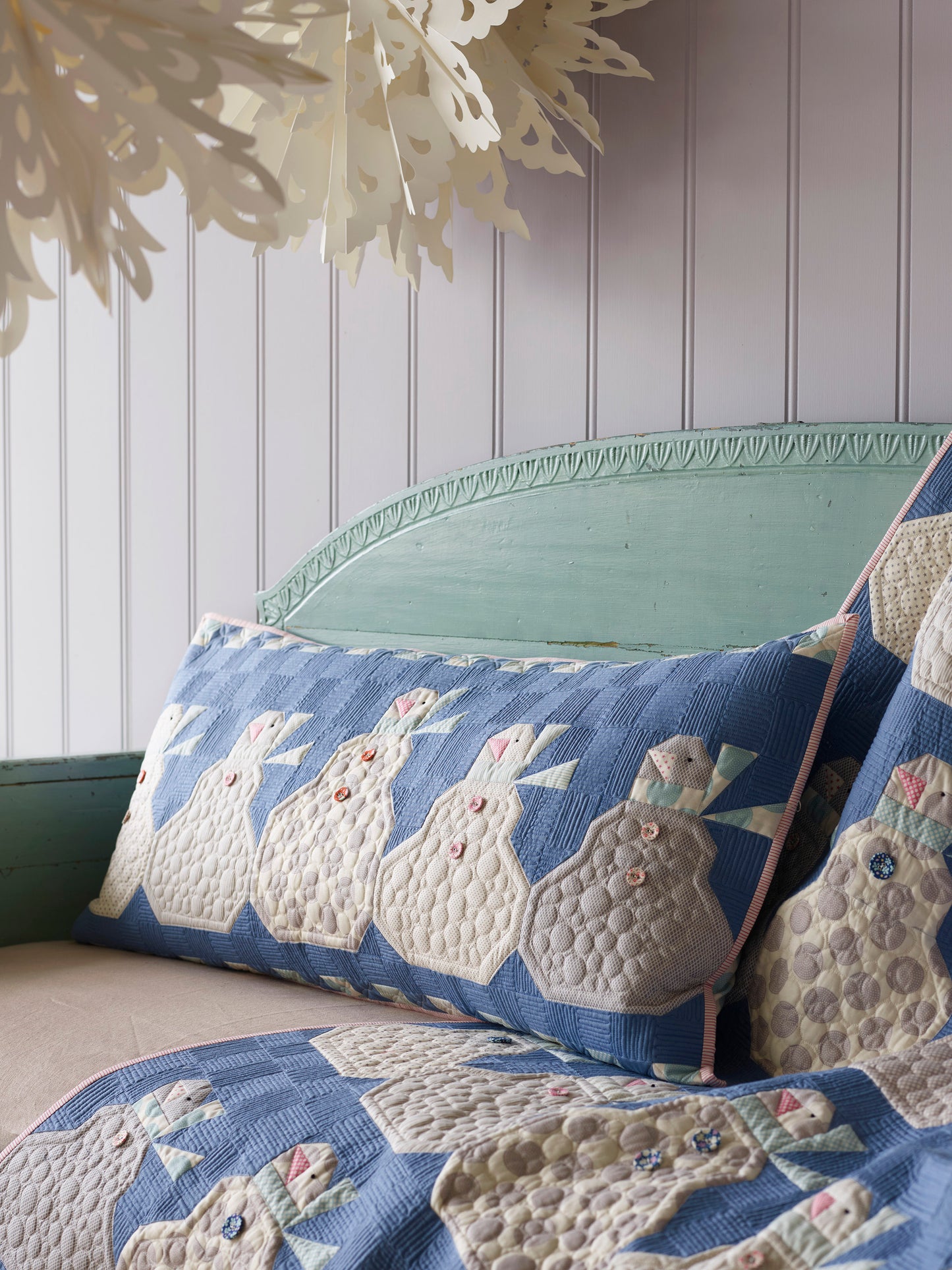 A quilted throw blanket and matching pillow featuring a blue and cream bunny pattern, styled on a distressed green bench against a white paneled wall.
Photo by Tildafabrics.com [image_link]
