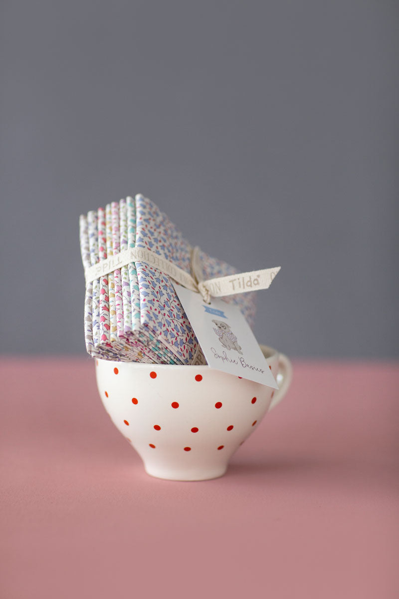 A small stack of patterned tea bags tied with a white ribbon and tag, displayed in a white ceramic teacup with red polka dots on a pink surface against a gray background.
Photo by Tildafabrics.com [additional_image_link]