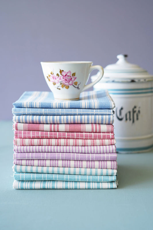 A stack of folded patterned quilt fat quarters in red, blue, green, purple and white stripes, with two matching ceramic teacups stacked on top, set against a split blue and dusty pink background.
Photo by Tildafabrics.com [image_link]