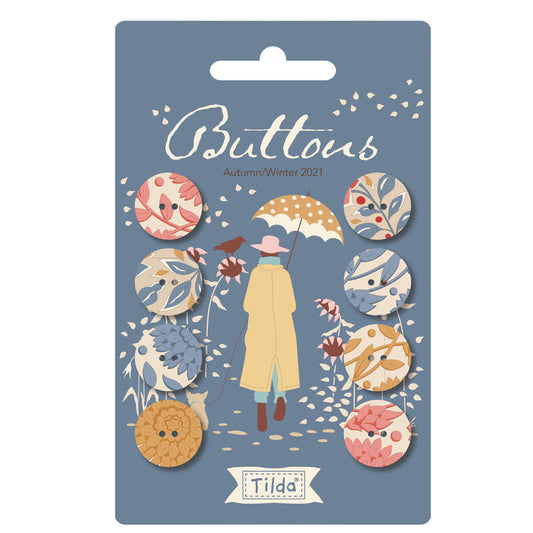 A package of Tilda brand buttons, labeled 'Butters Autumn/Winter 2021'. The packaging is slate blue with an illustration of a person in a yellow raincoat holding an umbrella, surrounded by eight circular buttons featuring various autumn and winter patterns like leaves, birds, and flowers.
Photo by Tildafabrics.com [image_link]