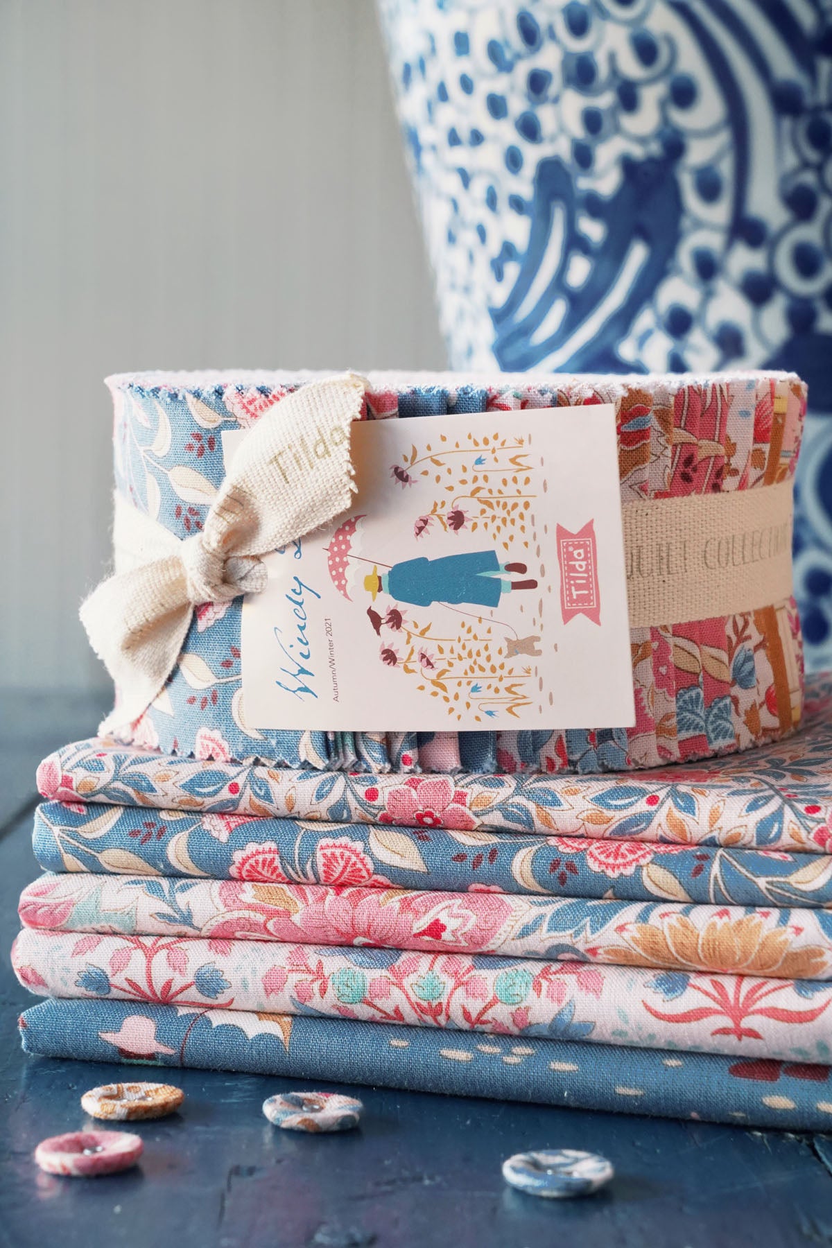 A stack of folded fabric bundles with floral patterns in blue, pink, and beige, tied with a cream ribbon and a paper tag that reads 'Windy Creek Fabrics Collection.' A blue-and-white patterned ceramic vase is blurred in the background, and a few buttons sit on a dark tabletop in the foreground.
Photo by Tildafabrics.com [additional_image_link]