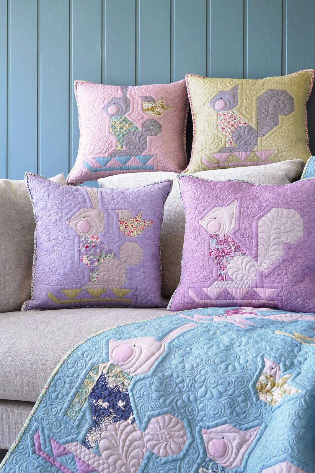 Four decorative throw pillows with quilted squirrels in pastel colors, arranged on a light beige sofa with a blue quilt draped over the front, set against a blue paneled wall.
Photo by Tildafabrics.com [additional_image_link]